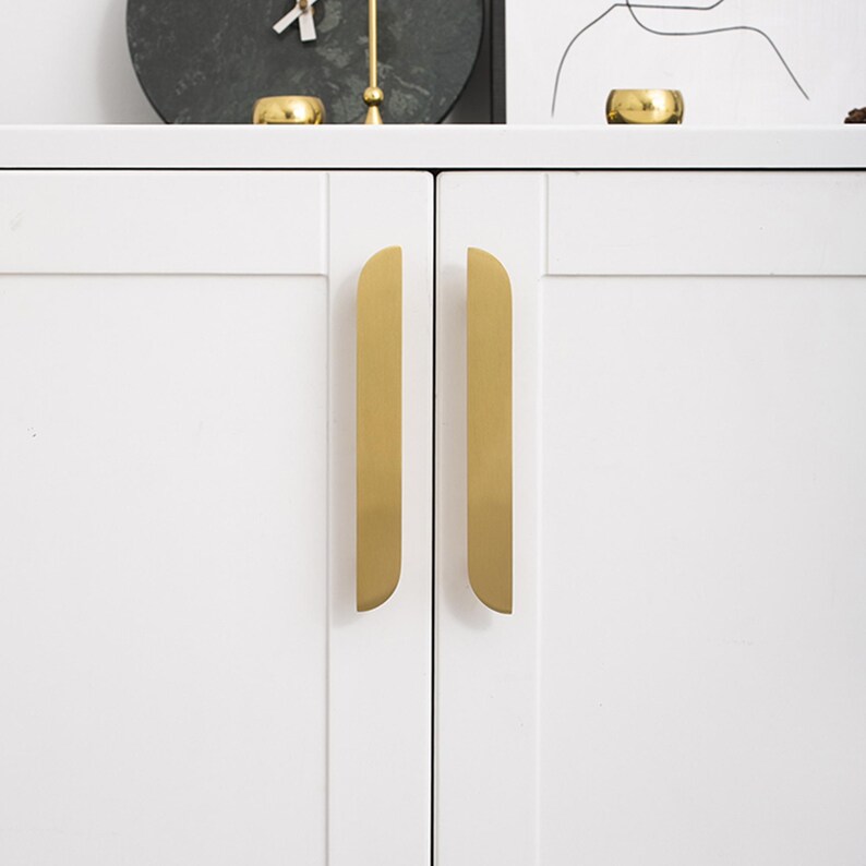 May include: Close-up of a white cabinet with two gold-coloured, vertical handles. The handles have a rounded top and are mounted on the doors. The cabinet is part of a larger piece of furniture, with decorative items on top.