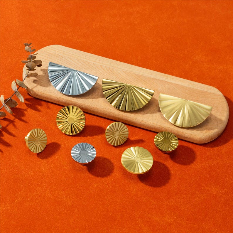 May include: A collection of decorative cabinet hardware in gold and silver tones. The hardware includes half-circle and circular designs with a radial pattern, displayed on a wooden cutting board against an orange background.