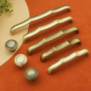 Może przedstawiać: Asortyment antycznych mosiężnych okuć do szafek, w tym kilka zakrzywionych uchwytów i trzy różne style okrągłych gałek. Okucia są prezentowane na czerwonopomarańczowej powierzchni. Uchwyty mają bambusowy design.