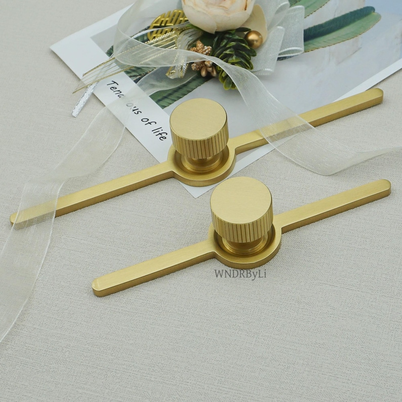 May include: Two gold cabinet pulls, each with a cylindrical, ridged knob and a rectangular handle. The pulls are on a light surface. The text "WNDRByLi" is visible. Ideal for drawers or cupboards.