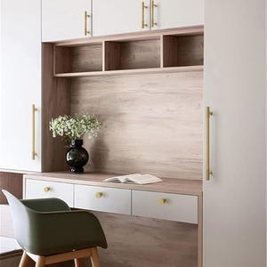 Puede incluir: Una moderna configuraci&oacute;n de oficina en casa con un escritorio con cajones blancos y herrajes dorados. Una silla verde est&aacute; frente al escritorio, y un jarr&oacute;n negro con flores blancas est&aacute; sobre el escritorio. El escritorio est&aacute; integrado en un armario blanco.