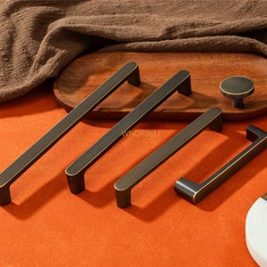 May include: A collection of bronze-toned cabinet hardware, including long bar pulls, a smaller pull, and a round knob. The hardware is displayed on an orange surface with a wooden tray and a brown cloth in the background.