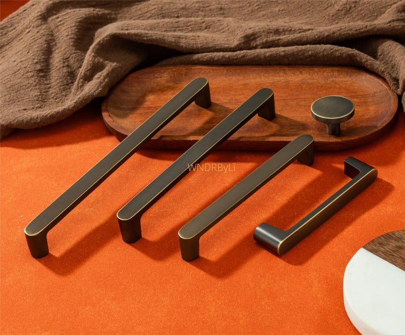 May include: A collection of bronze-toned cabinet hardware, including long bar pulls, a smaller pull, and a round knob. The hardware is displayed on an orange surface with a wooden tray and a brown cloth in the background.