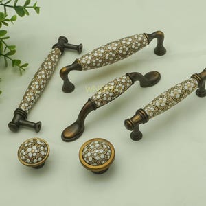 Könnte beinhalten: Eine Auswahl an dekorativen Schrankgriffen und -knöpfen. Die Griffe und Knöpfe weisen ein weißes Blumenmuster auf cremefarbenem Grund auf, mit bronzefarbenen Metallakzenten. Geeignet für Möbel und Schränke.