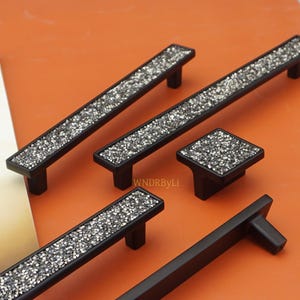 Peut inclure: Ensemble de quincaillerie de meubles noire avec des accents de cristaux argentés scintillants. L'ensemble comprend de longues barres de tirage et un bouton carré, tous avec un design moderne et élégant. Sur fond orange.