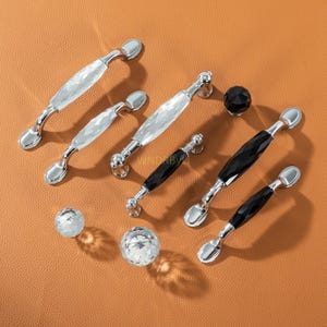 May include: A collection of cabinet hardware, including crystal and black faceted glass handles and knobs with silver-toned metal accents. The handles vary in size and shape, offering a range of design options for furniture and cabinetry. The hardware is displayed on a brown surface.