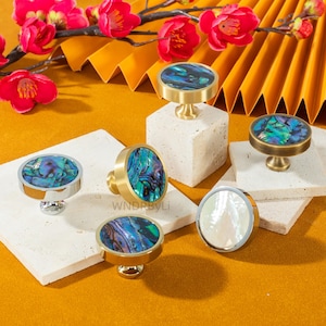 May include: A collection of decorative cabinet knobs with iridescent abalone shell centers. The knobs have brass, silver, and gold finishes. The knobs are displayed on a yellow surface with red flowers in the background.