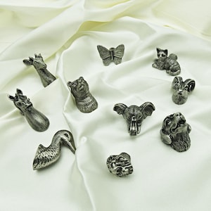 May include: A collection of small, silver-toned animal figurines, including a giraffe, bear, butterfly, cat, ram, elephant, lion, and bird. The figurines are arranged on a white, silky fabric, showcasing their detailed designs.