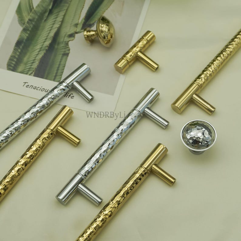 May include: An assortment of cabinet hardware, including silver and gold-toned T-bar handles and a faceted knob. The handles have a textured, hammered appearance. The image also shows a small gold knob. The text "Tenacious life" is visible in the background.