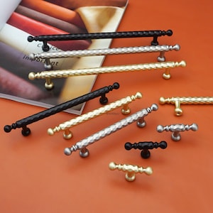 May include: An assortment of cabinet handles in various metallic finishes, including black, silver, and gold. The handles have a geometric diamond pattern and are displayed on an orange surface.