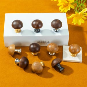 Könnte beinhalten: Eine Sammlung von pilzförmigen Holzschrankknöpfen in verschiedenen Holztönen. Einige Knöpfe haben goldene, silberne oder schwarze Metallsockel. Die Knöpfe werden auf einer weißen Oberfläche und einem orangefarbenen Hintergrund präsentiert. Der Text "WNDRByLi" ist sichtbar.