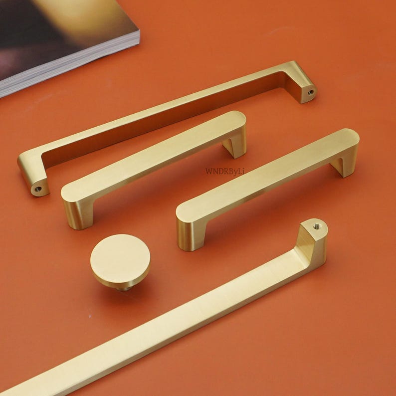 May include: A collection of gold-colored cabinet hardware, including various sizes of handles and a round knob. The hardware is arranged on an orange surface, showcasing different designs and lengths for diverse furniture applications.