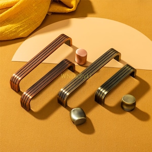 Może przedstawiać: Asortyment okuć do szafek, w tym uchwyty i gałki w odcieniach brązu i mosiądzu. Uchwyty mają zakrzywioną konstrukcję z wieloma równoległymi liniami. Gałki są cylindryczne i owalne. Ciepłe, neutralne tło.