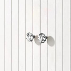 May include: Two silver cabinet knobs with a radial design are mounted on a white, vertically-paneled surface. The knobs have a circular shape with a series of radiating lines, creating a sunburst effect. The background is a clean, minimalist design.