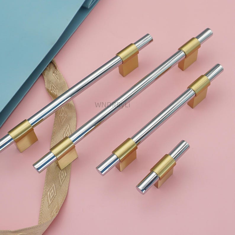 May include: A set of four cabinet pulls. The pulls have a brushed silver bar with gold-colored accents. The pulls are rectangular and have a modern design. The background is a light pink color.