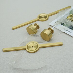May include: Gold-coloured cabinet hardware set, including two long bar pulls and two cylindrical knobs. The knobs have a ridged texture. The hardware is displayed on a light-coloured surface.