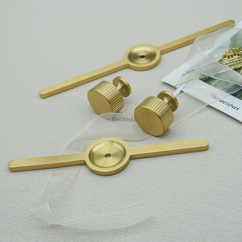 May include: Gold-coloured cabinet hardware set, including two long bar pulls and two cylindrical knobs. The knobs have a ridged texture. The hardware is displayed on a light-coloured surface.