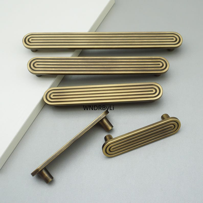 Op de afbeelding: Een verzameling antiek messing kastknoppen en handgrepen. De hardware heeft een gegroefd, rechthoekig ontwerp met afgeronde uiteinden. De handgrepen zijn in verschillende maten, geschikt voor laden en deuren. De afbeelding toont de hardware tegen een neutrale achtergrond.