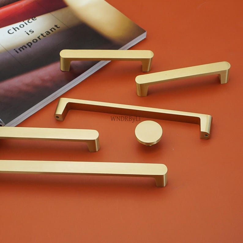 May include: A collection of gold-colored cabinet hardware, including various handle styles and a round knob, arranged on an orange surface. The handles are rectangular with rounded edges. The text "Choice is important" is visible on a book in the background.