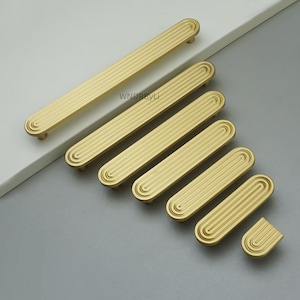 Op de afbeelding: Een verzameling goudkleurige kastgrepen in verschillende maten. Elke greep heeft een gegroefd, rechthoekig ontwerp met afgeronde uiteinden. De grepen zijn gerangschikt op een grijs en wit oppervlak, waarbij hun verschillende lengtes en stijlen worden getoond.