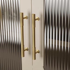 Puede incluir: Primer plano de dos tiradores dorados para puertas de armario. Los tiradores son verticales, cil&iacute;ndricos y tienen una textura acanalada. Las puertas del armario son de color beige claro y los paneles de vidrio tienen un patr&oacute;n acanalado vertical.