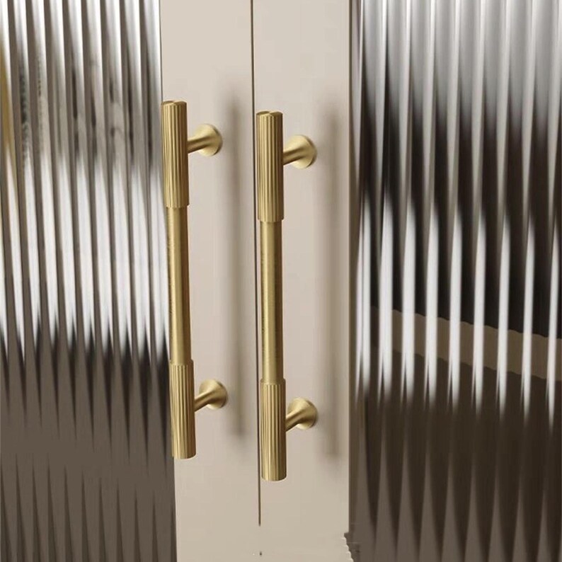 Puede incluir: Primer plano de dos tiradores dorados para puertas de armario. Los tiradores son verticales, cil&iacute;ndricos y tienen una textura acanalada. Las puertas del armario son de color beige claro y los paneles de vidrio tienen un patr&oacute;n acanalado vertical.