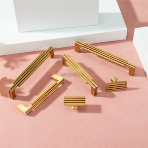 Puede incluir: Un conjunto de herrajes dorados para armarios, que incluyen tiradores y pomos. Los tiradores presentan un dise&ntilde;o lineal con tres l&iacute;neas paralelas, y los pomos tienen una est&eacute;tica similar. El hardware se muestra sobre una superficie rosa, con el texto "WNDRBYL" visible.