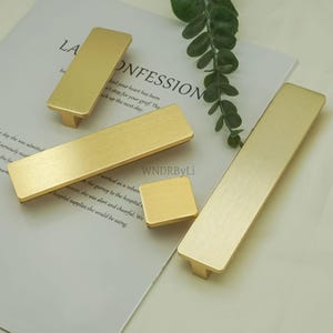 Może przedstawiać: Kolekcja złotych uchwytów i gałek do szafek w różnych prostokątnych kształtach. Okucia ułożone są na jasnej powierzchni z tekstem i gałązką zieleni. Uchwyty mają szczotkowane metalowe wykończenie.