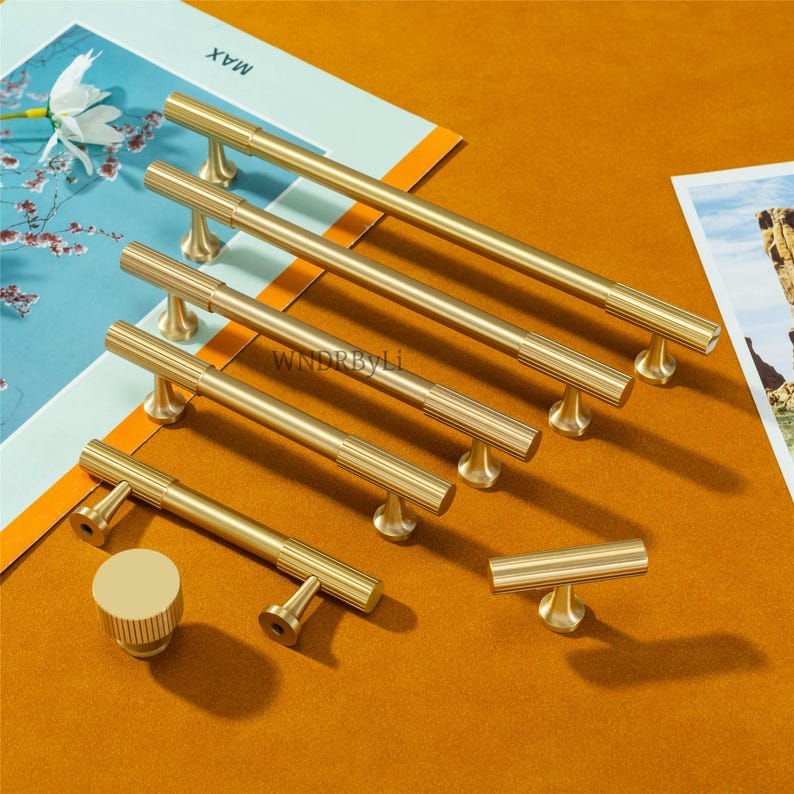 Puede incluir: Una colecci&oacute;n de herrajes dorados para armarios, que incluye varios tiradores de barra con detalles acanalados, un pomo redondo y un tirador en forma de T. Los herrajes est&aacute;n dispuestos sobre una superficie de color mostaza, con una impresi&oacute;n floral y la palabra "MAX" visible.