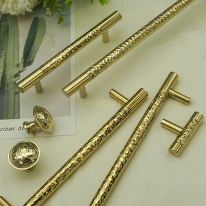 May include: Gold-toned cabinet hardware set featuring hammered texture. Includes various handle styles: long bar pulls, T-bar pulls, and round knobs. The hardware is displayed on a light background, showcasing the reflective quality of the metal.