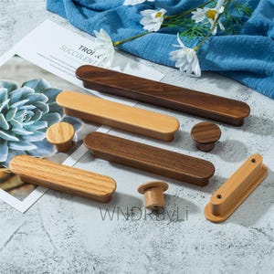 May include: A collection of wooden cabinet hardware, including long rectangular pulls and round knobs, in various shades of brown and tan. The hardware is displayed on a light gray surface with a blue cloth and white flowers in the background. The text "SUCCULENT PLA" is visible.