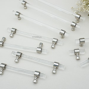 May include: Clear acrylic cabinet pulls with polished silver-tone hardware. The pulls have a cylindrical design and are arranged on a white surface. The text "WNDRByLi" is visible on the surface.