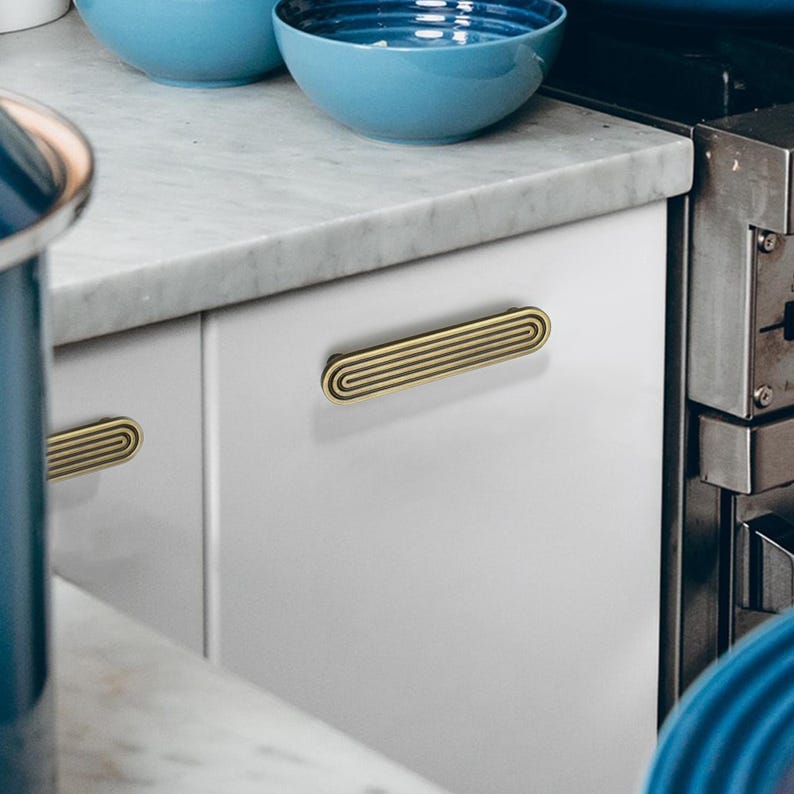 Op de afbeelding: Close-up van een witte keukenkast met twee goudkleurige, horizontale ladehandgrepen. De handgrepen hebben een gegroefd, ovaal ontwerp. Blauwe kommen zijn zichtbaar op het aanrecht. De afbeelding benadrukt keukenhardware en woondecoratie.