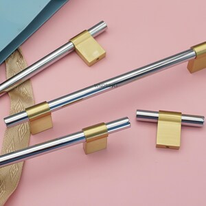 Op de afbeelding: Een collectie kastbeslag met een gepolijste zilveren staaf en goudkleurige vierkante uiteinden. Het beslag is gerangschikt op een roze oppervlak, wat een moderne design esthetiek laat zien. De afbeelding benadrukt het contrast tussen zilver en goud.