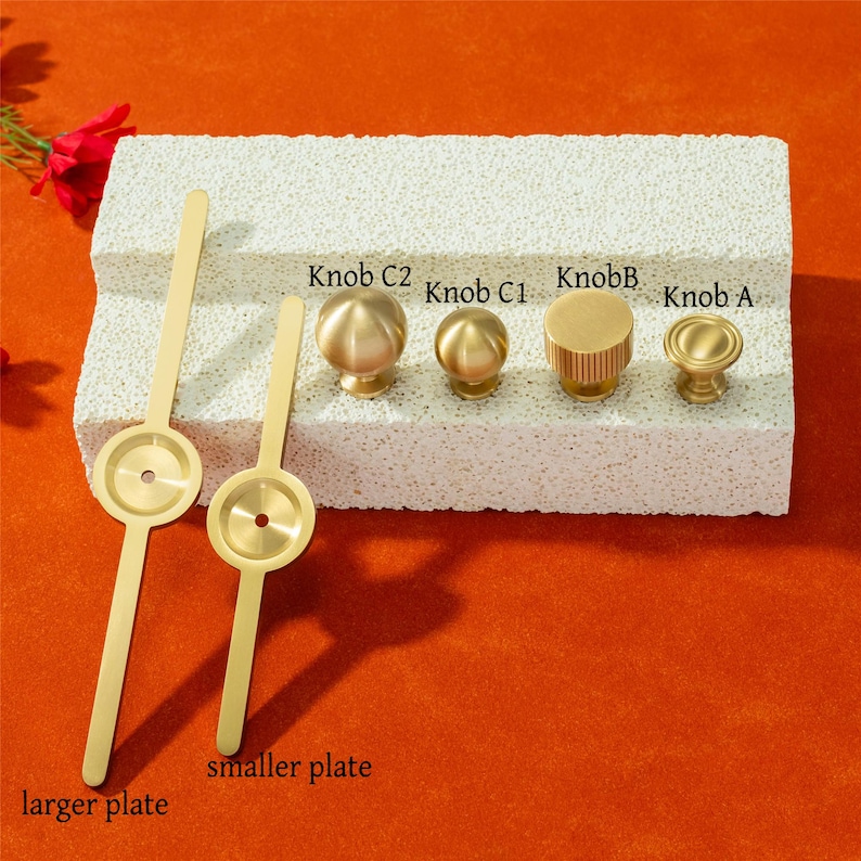 May include: A selection of brass cabinet knobs and plates presented on a textured white surface against an orange backdrop. The knobs showcase diverse shapes, such as spheres and cylinders. The plates are labelled "larger plate" and "smaller plate".