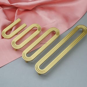 May include: Four gold-colored cabinet pulls with a grooved, elongated oval design. The pulls are arranged on a pink and gray surface, showcasing their hardware design. The pulls are made of metal and have a modern aesthetic.