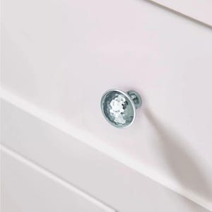 May include: A close-up of a white cabinet with a single, round, silver cabinet knob. The knob has a textured, faceted surface, reflecting light. The cabinet appears to be painted white.