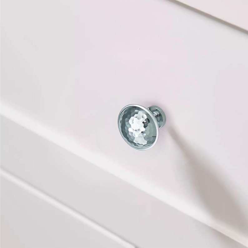 May include: A close-up of a white cabinet with a single, round, silver cabinet knob. The knob has a textured, faceted surface, reflecting light. The cabinet appears to be painted white.