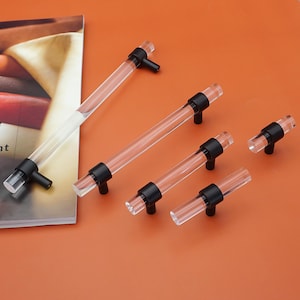 May include: A set of clear acrylic cabinet pulls with black metal detailing. The cylindrical pulls are bar-shaped and are displayed on an orange surface. These are designed for furniture and home decor.