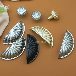 May include: A collection of decorative cabinet hardware in various metallic finishes. The assortment includes silver and gold knobs, along with silver, gold, and black half-moon shaped pulls. The hardware is displayed on a brown surface.