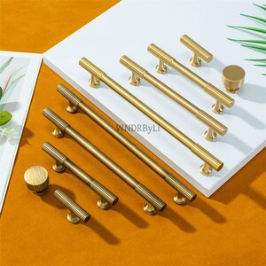 Puede incluir: Una colecci&oacute;n de herrajes dorados para gabinetes, que incluye varios tama&ntilde;os de tiradores de barra, perillas en T y una perilla redonda. Los herrajes presentan un dise&ntilde;o acanalado en la barra central y la perilla, con una base lisa y redondeada. Los art&iacute;culos est&aacute;n dispuestos sobre una superficie blanca.