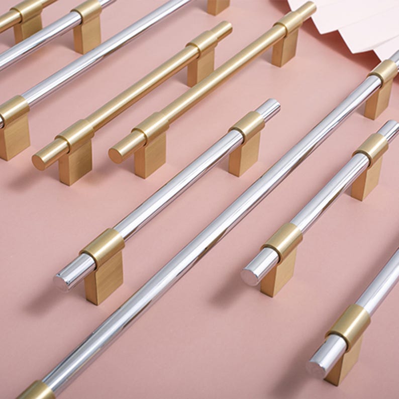 Op de afbeelding: Kast hardware met gouden en zilveren stanggrepen. De cilindrische stanggrepen hebben gouden accenten en zijn gemonteerd op goudkleurige rechthoekige bases. Opstelling op een lichtroze ondergrond.