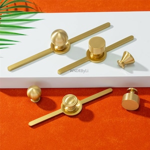 May include: A selection of gold-coloured cabinet hardware, including knobs and pulls. The hardware features various designs, such as spherical knobs, cylindrical knobs, and pulls with a central knob and a bar extending on either side. The text "WNDRByLi" is visible.