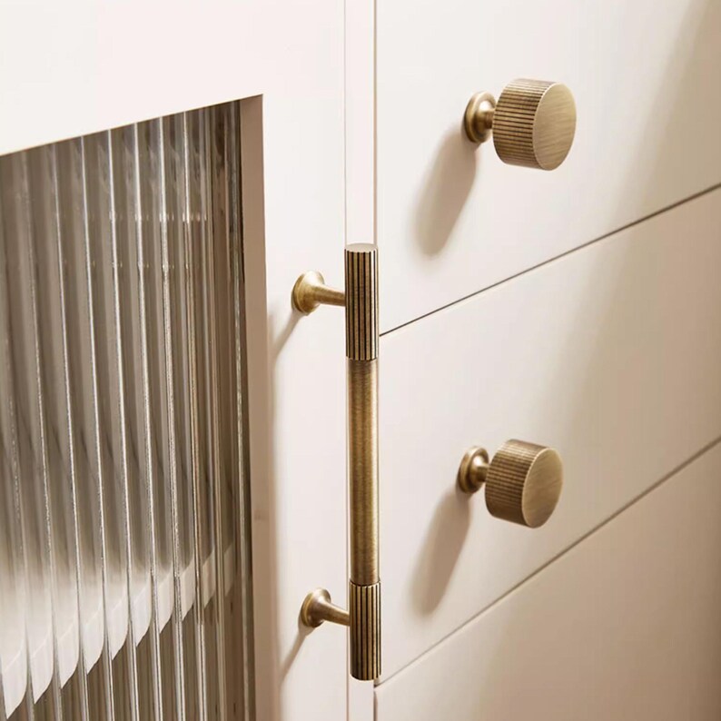 Puede incluir: Primer plano de herrajes de armario con dos pomos redondos y un tirador largo en acabado lat&oacute;n. Los pomos y el tirador tienen una textura acanalada. Las puertas del armario son blancas y se ve un panel de vidrio con ranuras verticales.