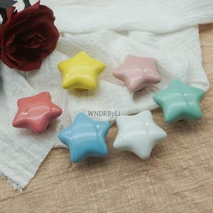 May include: Seven star-shaped ceramic knobs in pastel shades of yellow, pink, coral, mint green, light blue, and white. The knobs are displayed on a white fabric. The text "WNDRByLi" is visible.