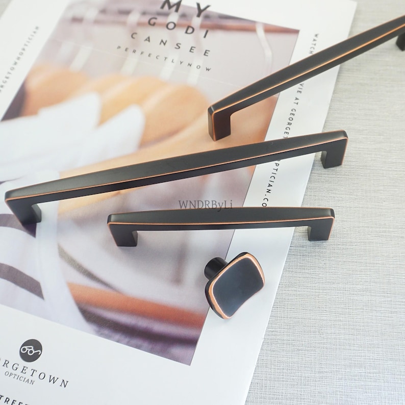 May include: A selection of dark bronze cabinet handles and a knob. The rectangular bar pulls and square knob have a modern design with a copper-coloured edge. The text "MY GOD I CAN SEE PERFECTLY NOW" is visible in the background.