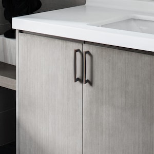 May include: A close-up of a bathroom cabinet featuring two bronze-coloured handles. The cabinet doors are light grey with a vertical wood grain pattern. The handles are rectangular with a slight curve and a decorative bottom.