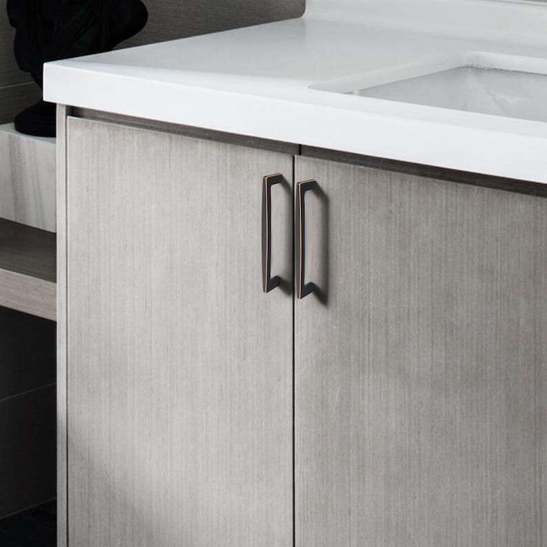 May include: A close-up of a bathroom cabinet featuring two bronze-coloured handles. The cabinet doors are light grey with a vertical wood grain pattern. The handles are rectangular with a slight curve and a decorative bottom.
