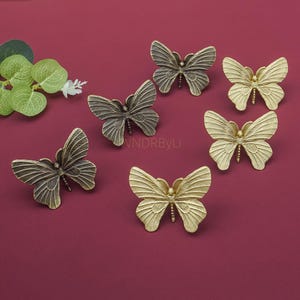 Könnte beinhalten: Sechs schmetterlingsförmige Möbelknöpfe in verschiedenen Gold- und Bronzetönen. Sie sind auf einer bordeauxfarbenen Oberfläche angeordnet, mit grünen Blättern oben links. Jeder Schmetterling hat detaillierte Flügelmuster.
