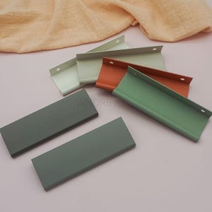 May include: A collection of colorful cabinet pulls in various shades of green, orange, and gray. The pulls are rectangular with a curved handle and two pre-drilled holes for mounting. The background is a soft pink with a peach-colored fabric.